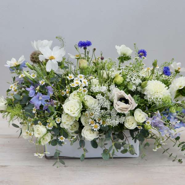 Low white ceramic centerpiece with white, blue, and purple flowers in a loose garden style
