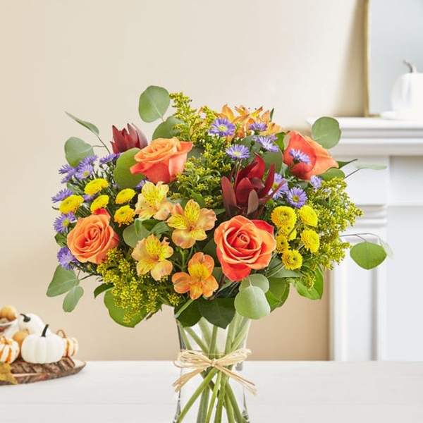 Mixed bouquet of orange roses, purple daisies, and yellow flowers in a glass vase