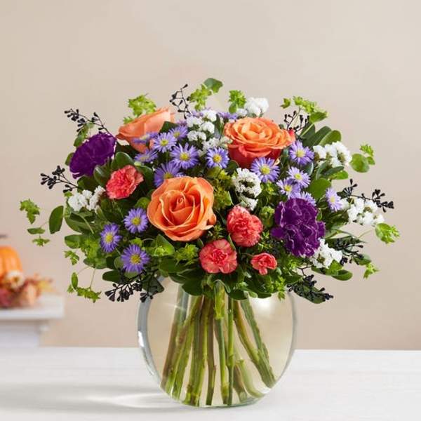 Mixed bouquet of orange roses, purple asters, and carnations in a glass vase