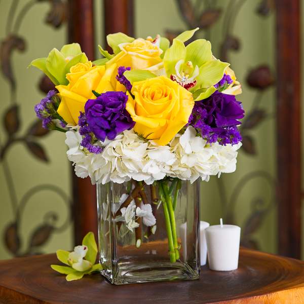 Yellow roses and purple flowers in a clear glass vase with white blooms