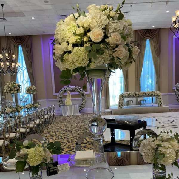 Tall white rose and hydrangea centerpiece in a clear glass vase on a mirrored table.