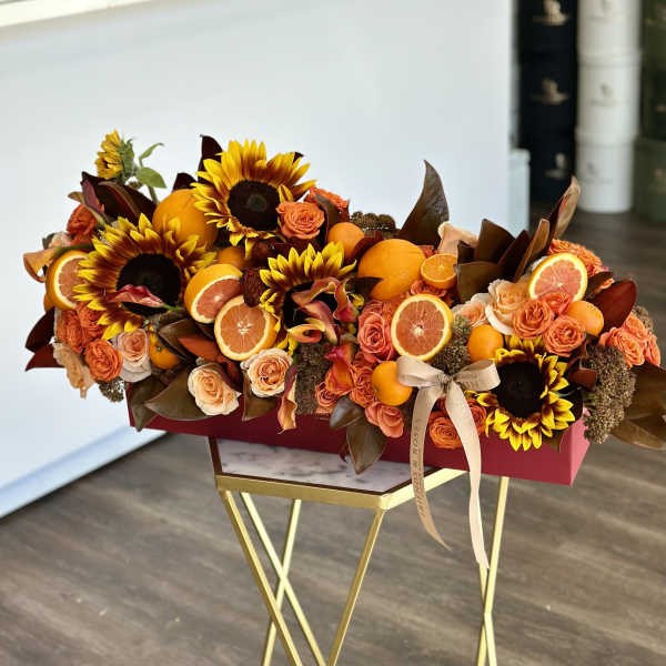 Orange roses and sunflowers arranged with sliced citrus in a red box.