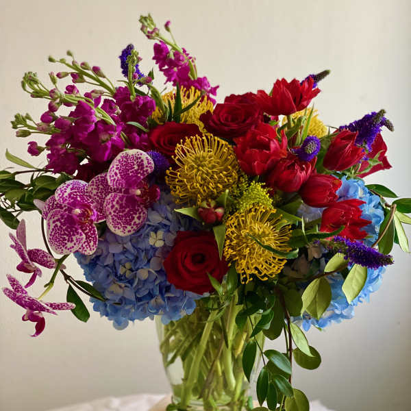 Tall mixed arrangement of red roses, yellow pincushion protea, blue hydrangeas, and spotted orchids in a clear glass vase