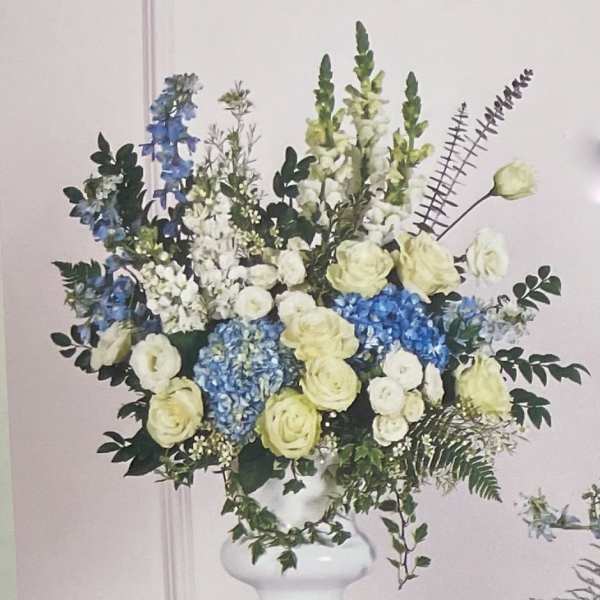 Large floral arrangement in a white pedestal vase with blue, white, and pale yellow blooms