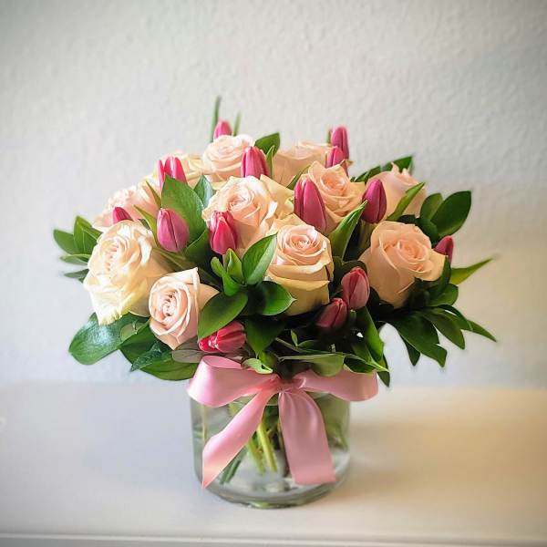 Blush roses and pink tulips arranged in a clear glass vase with a pink ribbon.