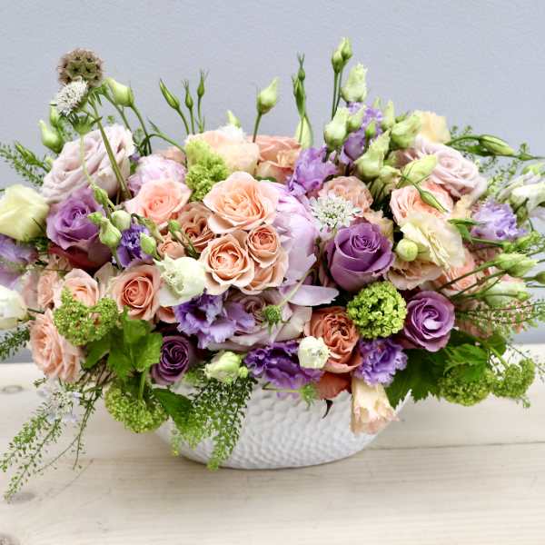 Low centerpiece with peach and lavender roses, lisianthus and green blooms in a white oval vase