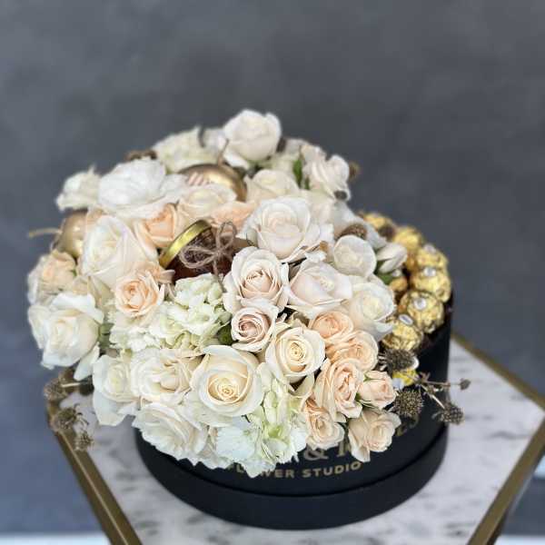 Cream and blush roses arranged in a round black box with gold chocolates
