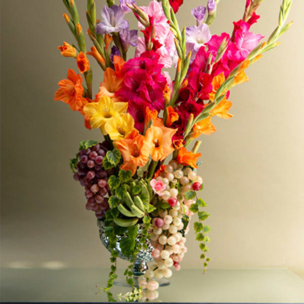 Tall arrangement of colorful gladiolus with grape clusters in a silver vase
