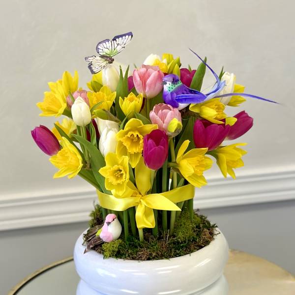 Bright arrangement of yellow daffodils and multicolor tulips in a white pot with decorative birds and butterfly