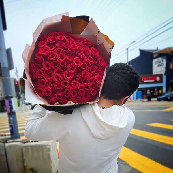 Large armful bouquet of tightly packed red roses wrapped in kraft paper, carried on a person's shoulder