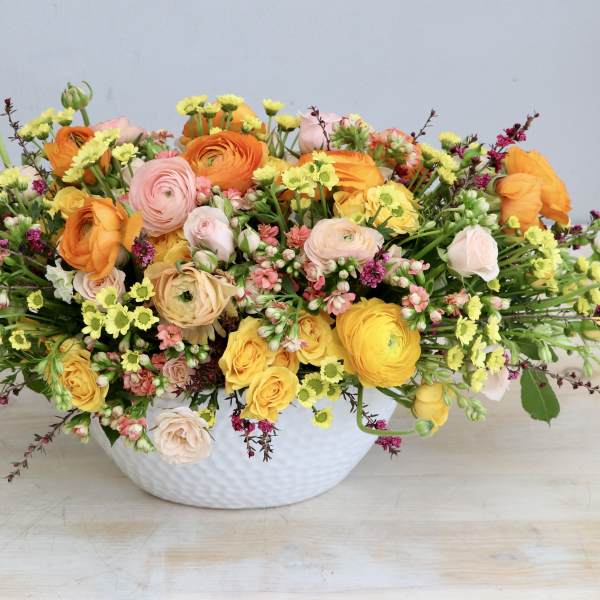Low centerpiece of yellow, orange, and blush flowers arranged in a white ceramic bowl