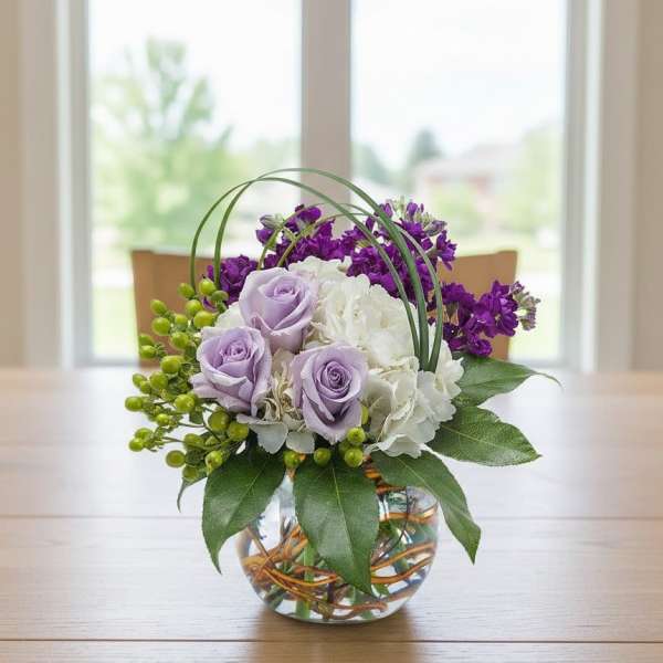 Round glass vase arrangement with lavender roses, white hydrangea, and purple flowers with green berries