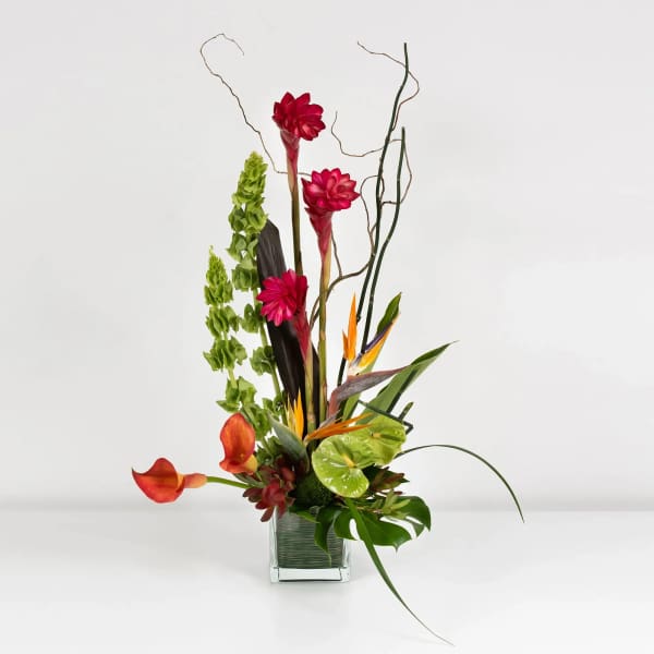 Tall tropical arrangement with red flowers in a clear square vase