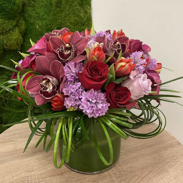 Low arrangement of red roses, burgundy orchids, and lavender flowers in a clear glass cylinder vase
