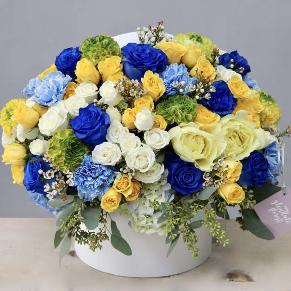 Round hatbox arrangement of yellow, blue, and white roses, carnations, and hydrangeas with a gift tag