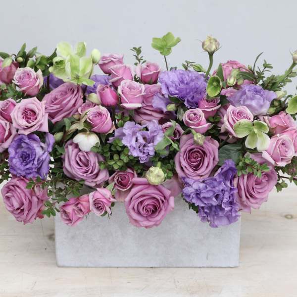 Low rectangular arrangement of pink and lavender roses with purple blooms in a white container