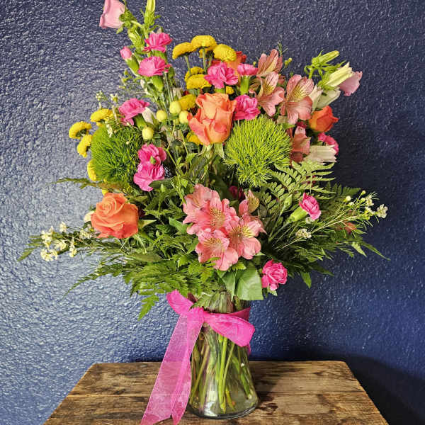 Mixed bouquet of pink, orange, and yellow flowers in a glass vase with a pink ribbon
