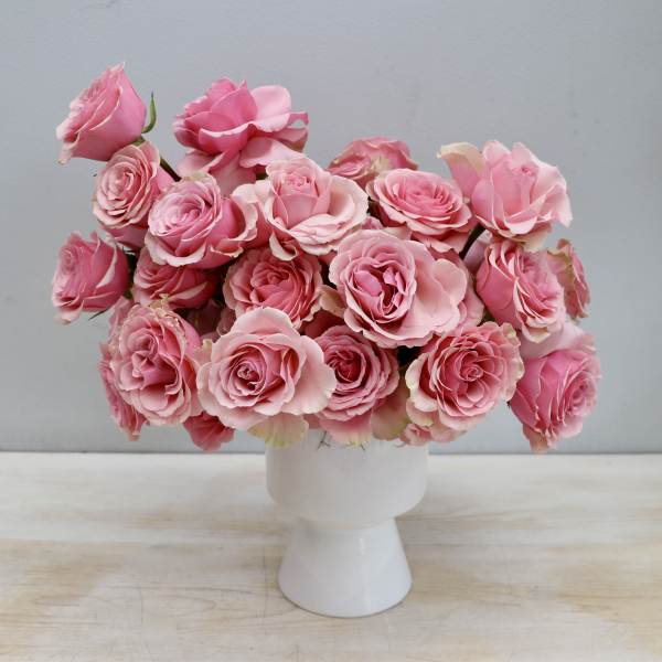 Low arrangement of pink roses in a white pedestal vase on a light wood surface