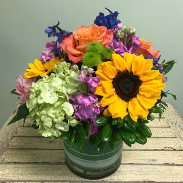 Mixed bouquet with a sunflower, roses, hydrangea, and purple blooms in a glass vase