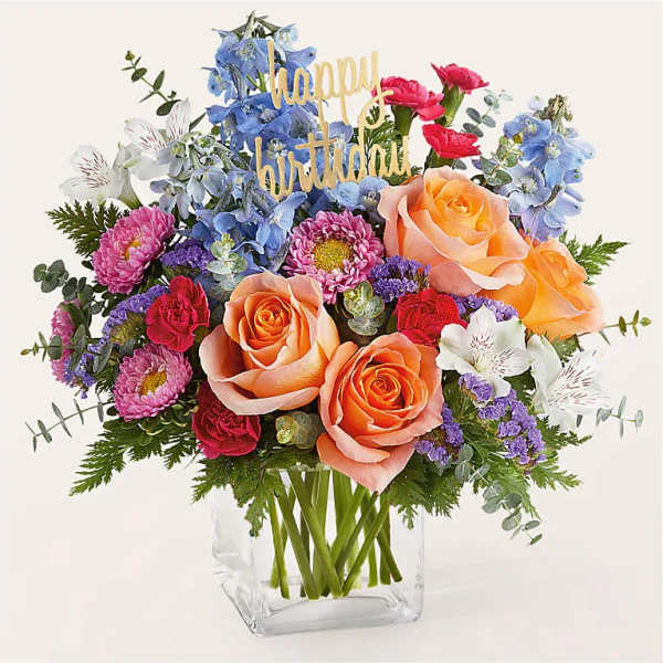 Mixed bouquet of roses, daisies, and blue flowers in a clear vase with a birthday sign