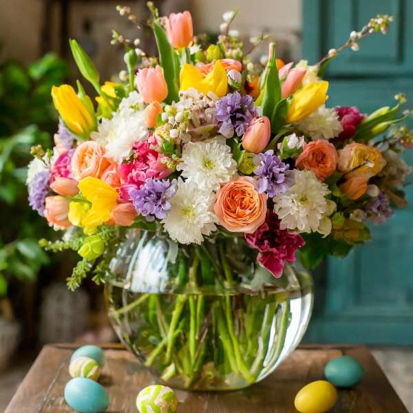 Colorful spring bouquet in a clear glass vase with painted eggs on a wooden table
