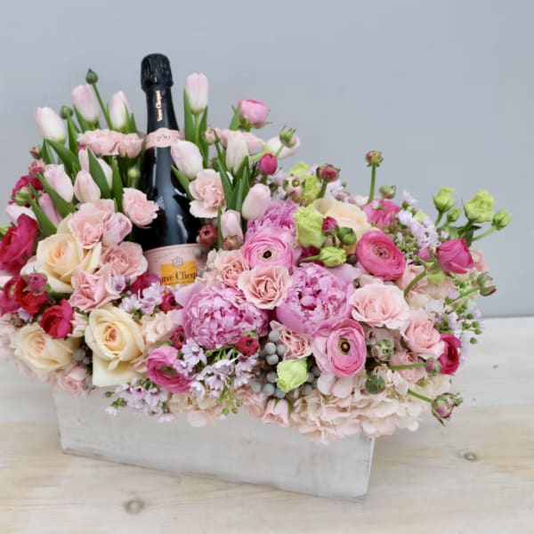 Rectangular pink and peach flower arrangement with tulips, roses, and peonies surrounding a champagne bottle.