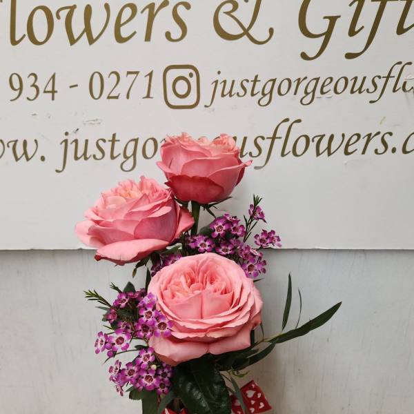 Pink roses and purple filler flowers in a clear glass vase with a red heart ribbon.