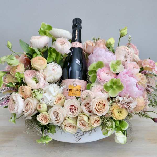 Low pastel flower arrangement in a white bowl with a pink champagne bottle in the center