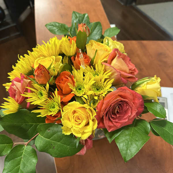 Low arrangement of yellow and orange roses and yellow mums in a clear glass vase on a wooden table.