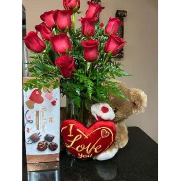 Tall arrangement of red roses in a clear vase with a teddy bear and chocolate box