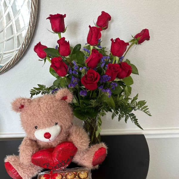 Red rose bouquet in a glass vase with purple blooms, a plush bear with red heart, and a box of chocolates.