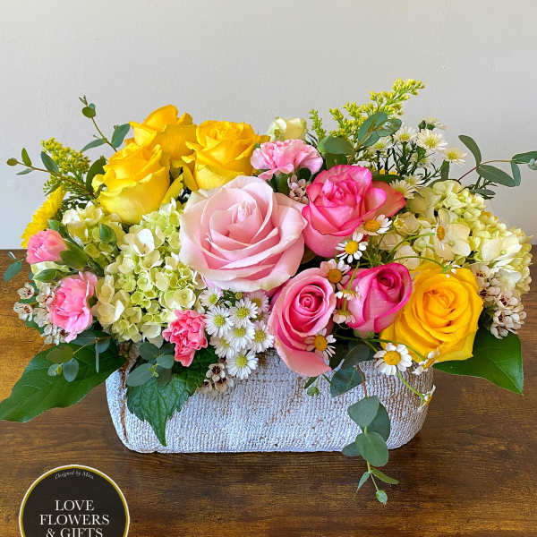 Low arrangement of pink and yellow roses with hydrangeas and daisies in a rectangular white container
