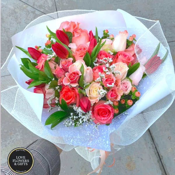 Handheld bouquet of pink roses and red tulips wrapped in white paper and mesh.