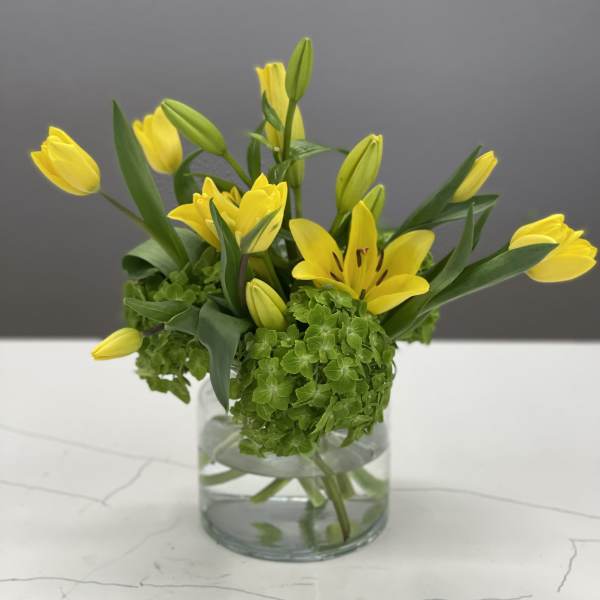 Yellow tulips and lilies arranged in a clear glass vase with green hydrangea