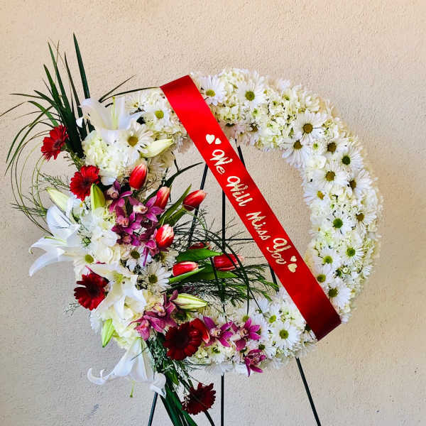 Heart-shaped funeral wreath with white daisies, lilies, and red ribbon on a stand