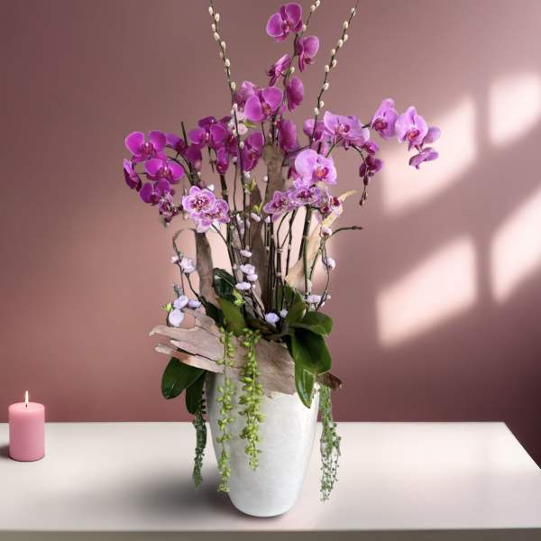 Purple orchid arrangement in a white vase with a pink candle beside it