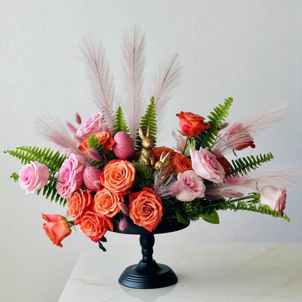 Pink and orange rose arrangement in a black pedestal vase with decorative eggs and bunny figurines