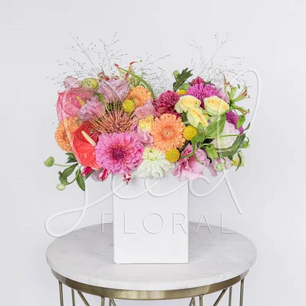 Bright mixed arrangement of pink, orange, and yellow flowers in a white square vase.