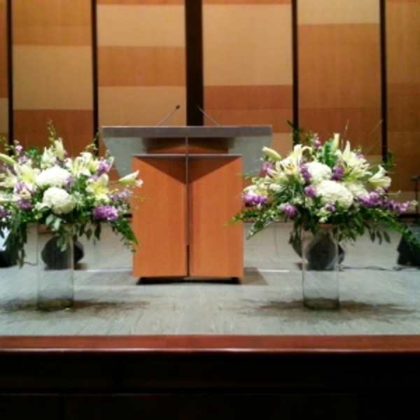 Two large white and purple floral arrangements in tall clear vases flanking a wooden podium