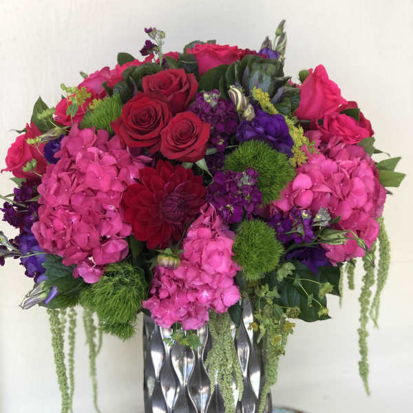 Tall arrangement of hot pink hydrangeas, red roses, and purple blooms in a silver patterned vase