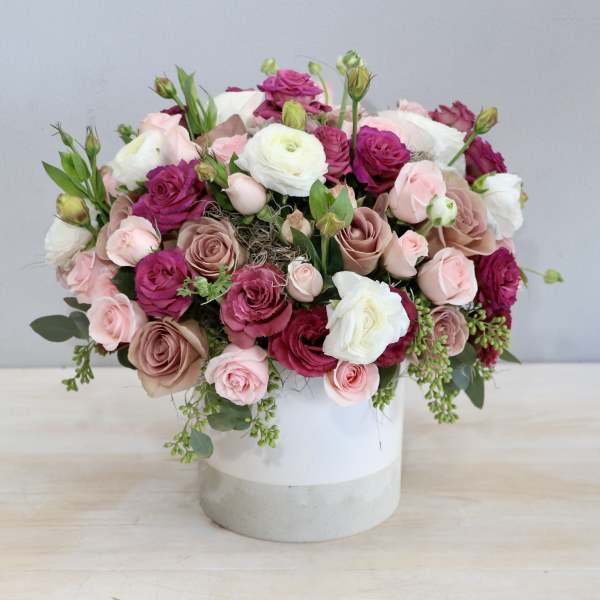 Low arrangement of pink, mauve, and white roses and ranunculus in a white cylinder vase