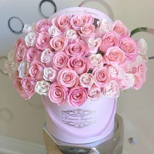 Round hatbox filled with tightly packed pink and white roses on a reflective stand.