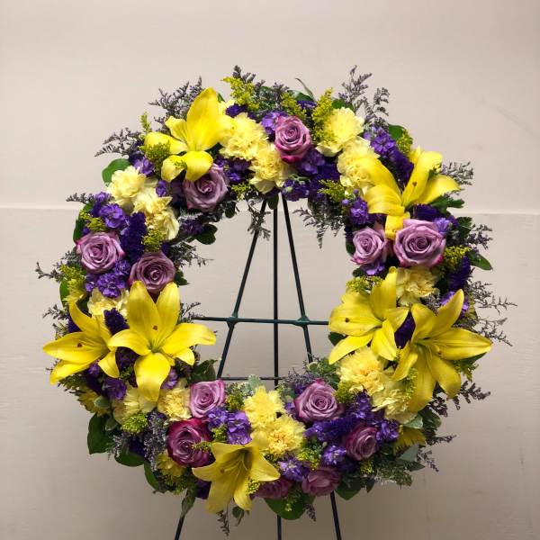 Standing wreath of yellow lilies, pale yellow blooms, and lavender and purple flowers on an easel