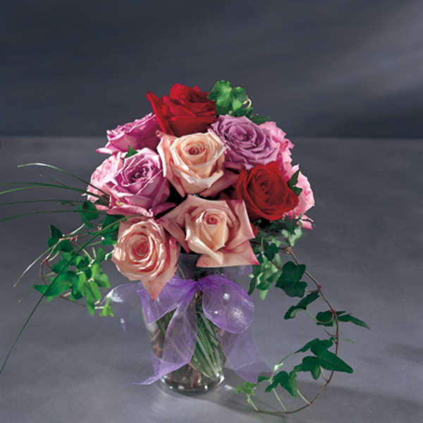 Bouquet of red, pink, and peach roses in a glass vase with a purple ribbon