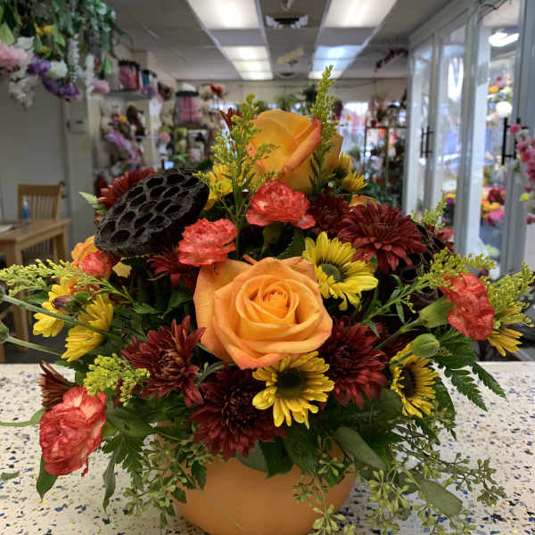 Orange rose arrangement with yellow and burgundy flowers in a pumpkin-shaped container