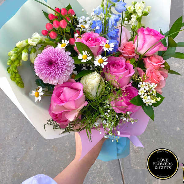 Hand-tied bouquet of pink roses, blue delphinium, white snapdragons, and mixed blooms wrapped in pastel paper