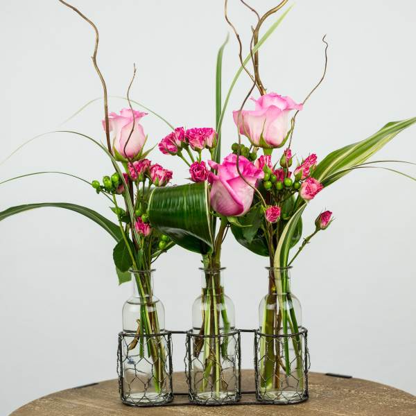 Three small glass vases hold pink roses and carnations with tall decorative branches.