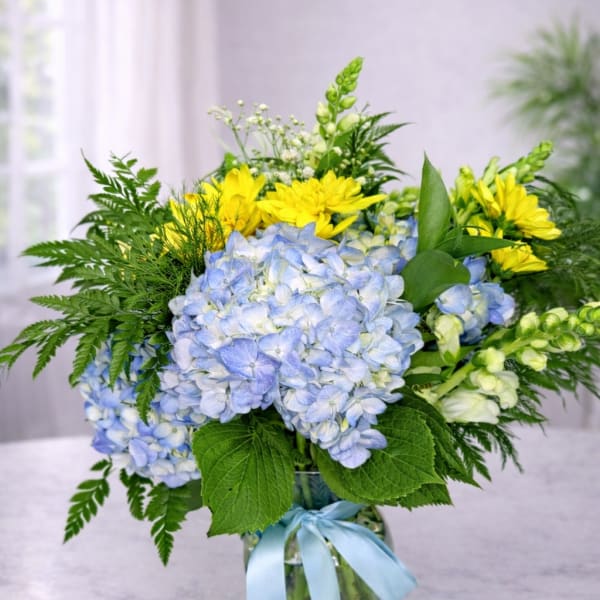 Blue hydrangeas and yellow daisies in a glass vase with a blue ribbon.