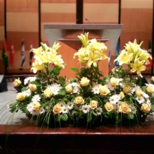 Low yellow lily, rose, and white daisy arrangement in a long container in front of a podium