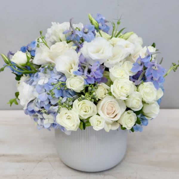 Low white ceramic vase with white roses, blue hydrangeas, and light blue flowers in a rounded arrangement